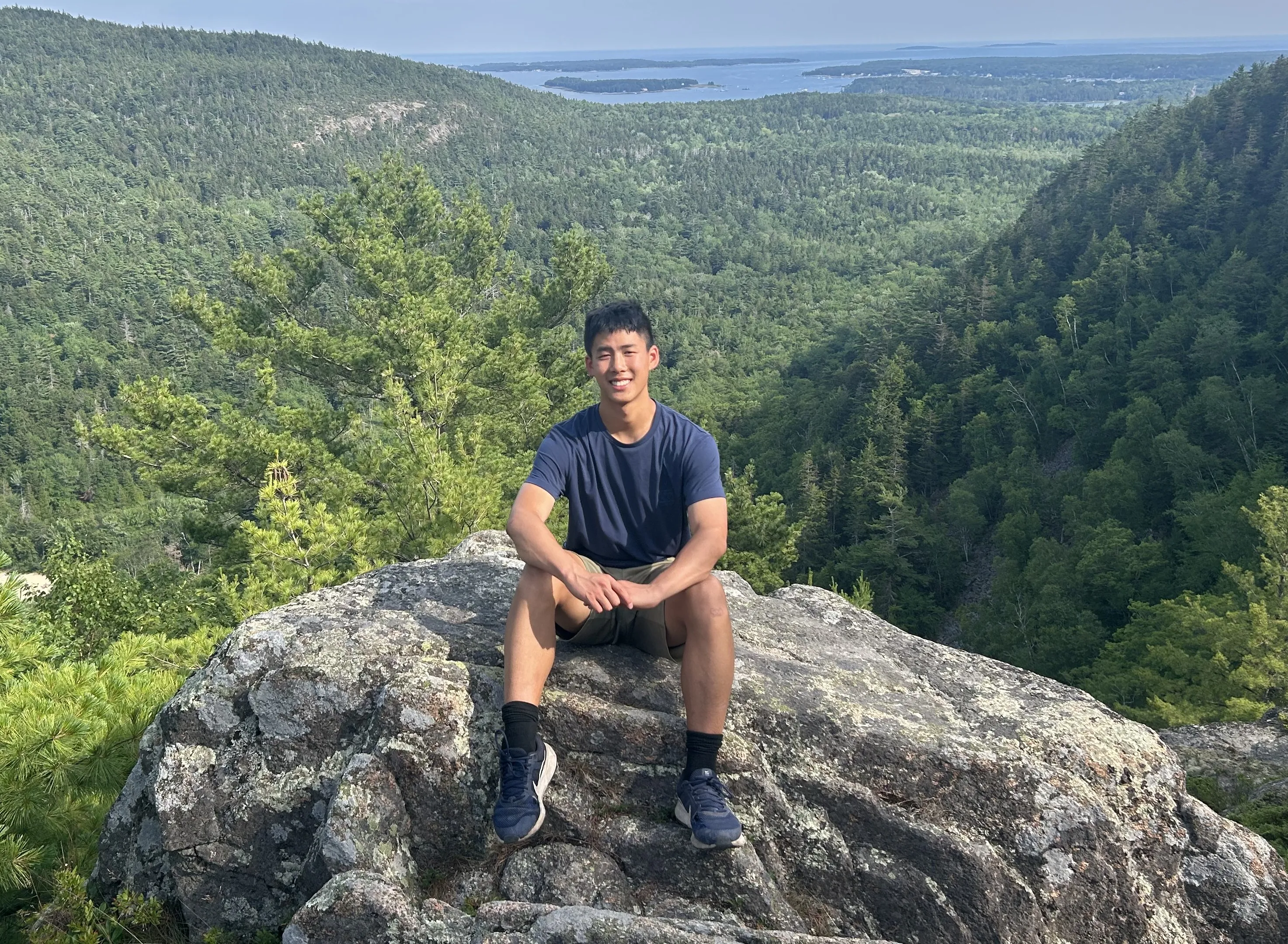 Kevin Chen sitting on a granite outcrop in Acadia National Park, a forested valley below him and the bay and its islands on the horizon behind.